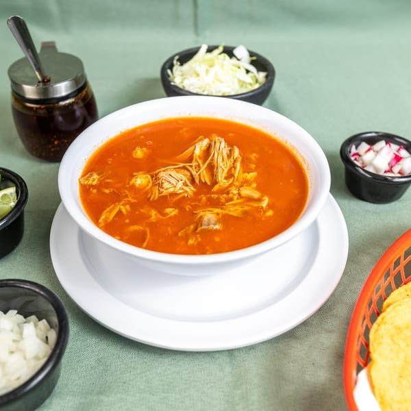 Red shredded meat soup with lime, onions, cabbage, radishes, chili oil, and tortillas on a green table.