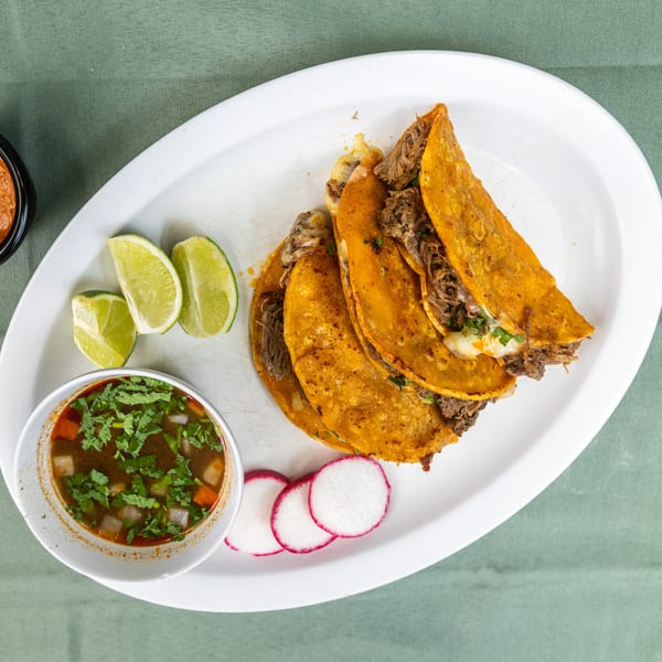 Birria tacos with consommé, lime wedges, radish slices, and red sauce on a white plate.