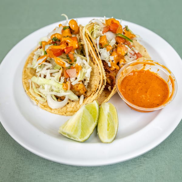 Two tacos topped with shredded cabbage, diced tomatoes, and orange sauce, served with lime wedges and a side of sauce.