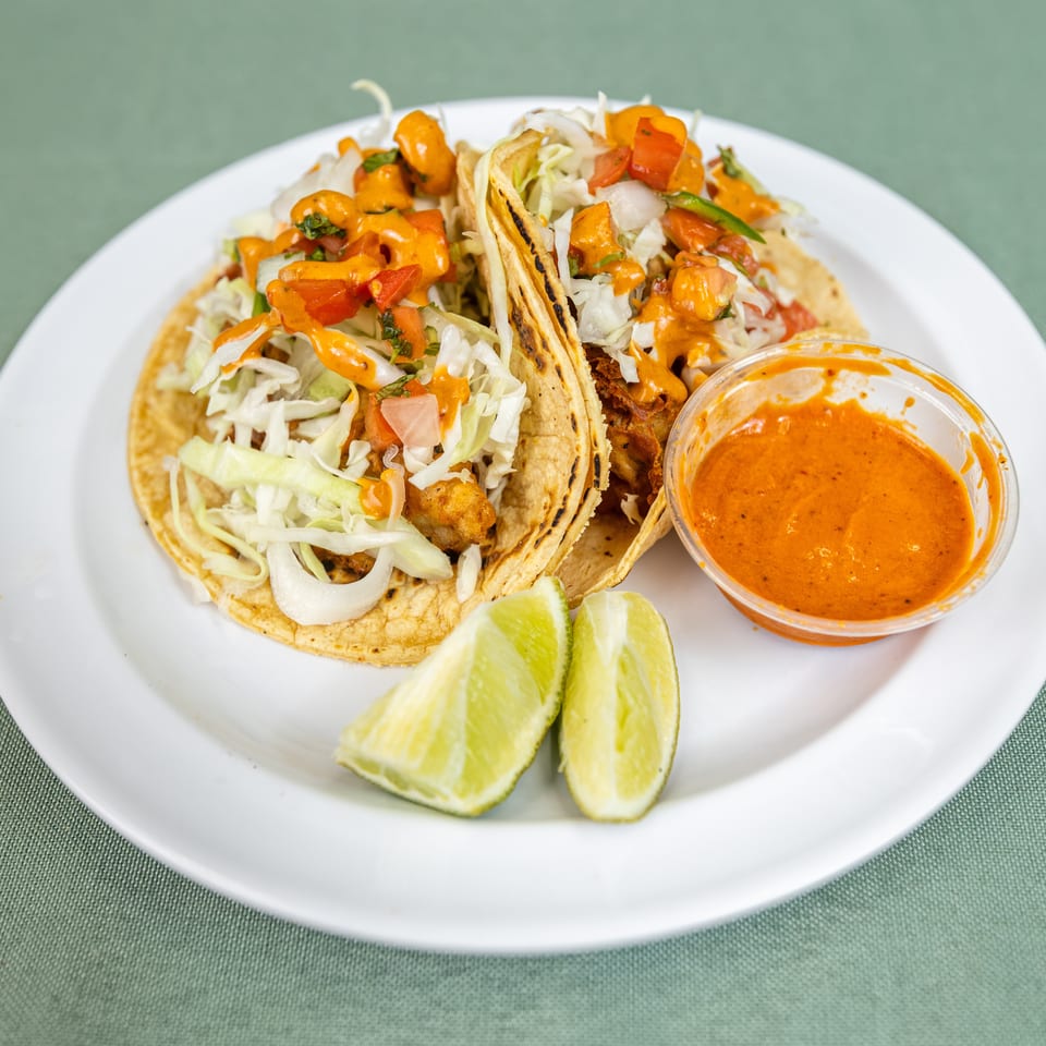 Two tacos topped with shredded cabbage, diced tomatoes, and orange sauce, served with lime wedges and a side of sauce.