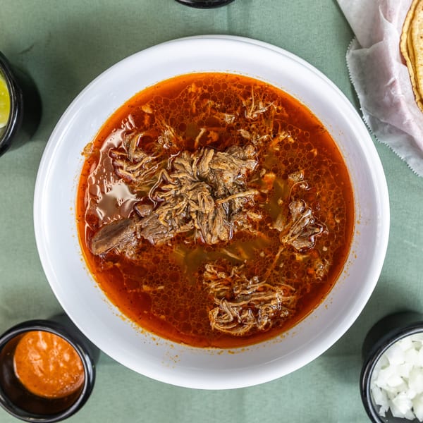 Hearty bowl of red birria soup with shredded meat, served with limes, onions, salsa, and tortillas.