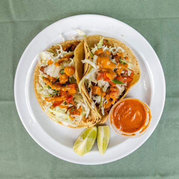 Two loaded tacos with shredded cabbage, diced tomatoes, and spicy orange sauce, plus lime wedges.