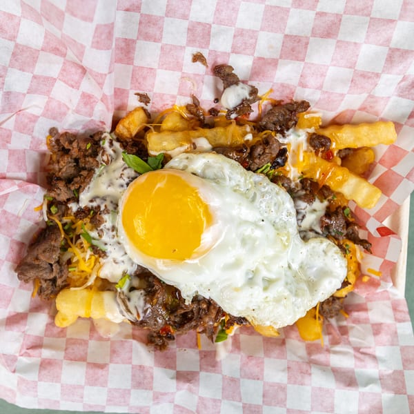 Loaded fries with ground meat, shredded cheese, creamy sauce, and a fried egg in a checkered paper.