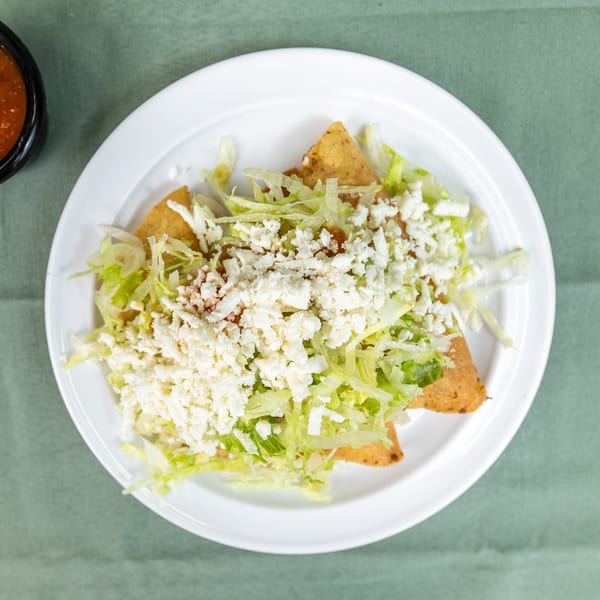 A white plate of golden taquitos covered in lettuce and white cheese, with a side of salsa.