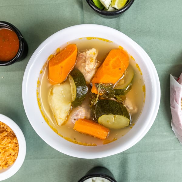 Chicken soup with carrots, zucchini, rice, salsa, limes, and tortillas on a green tablecloth.