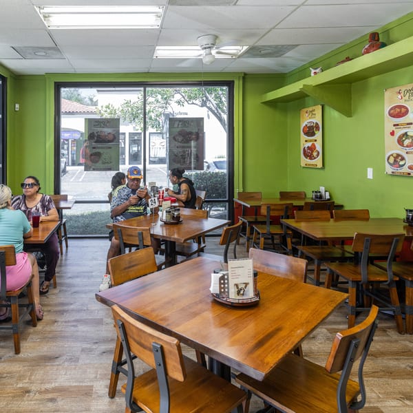 Diners seated at wooden tables in a casual Doña Elena Pozoleria restaurant with green walls and large windows.