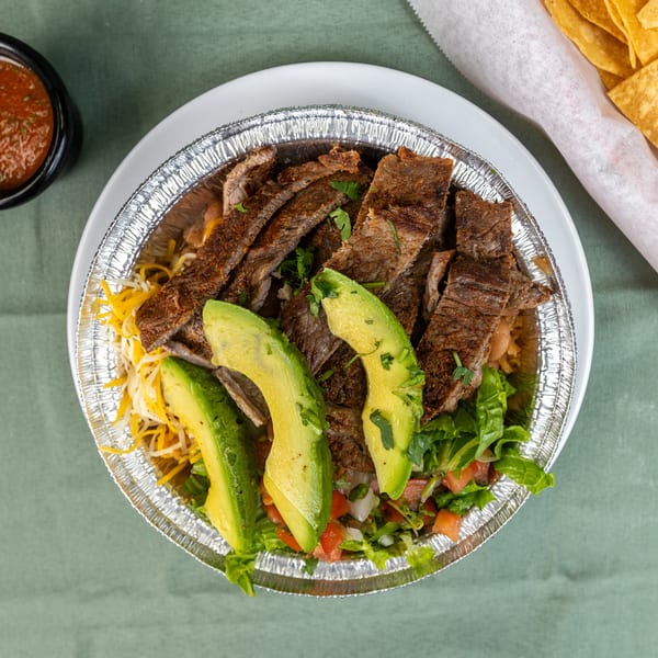 Sliced steak bowl with avocado, lettuce, cheese, salsa, and tortilla chips.