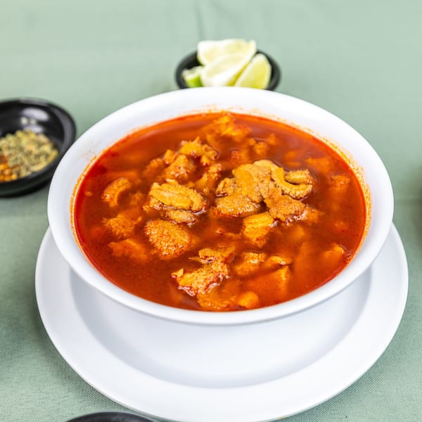 Red menudo soup with tripe in a white bowl, served with lime and spices.